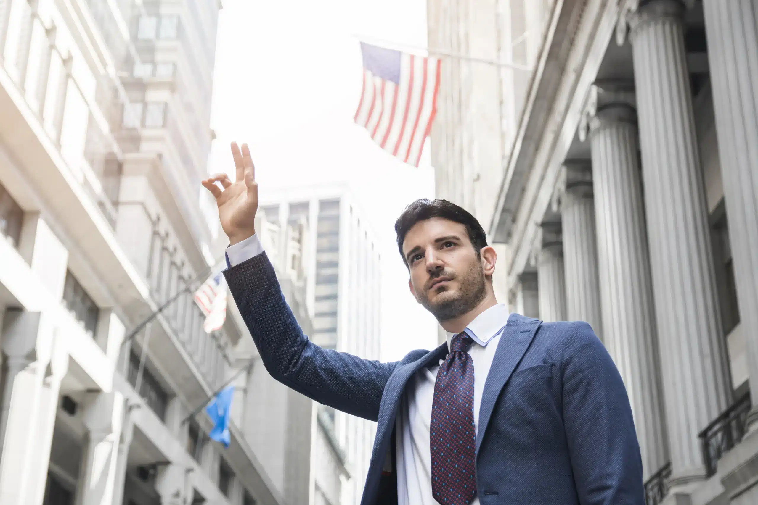 A man in a business suit gestures confidently while standing on a city street with U.S. flags and government-style buildings in the background.